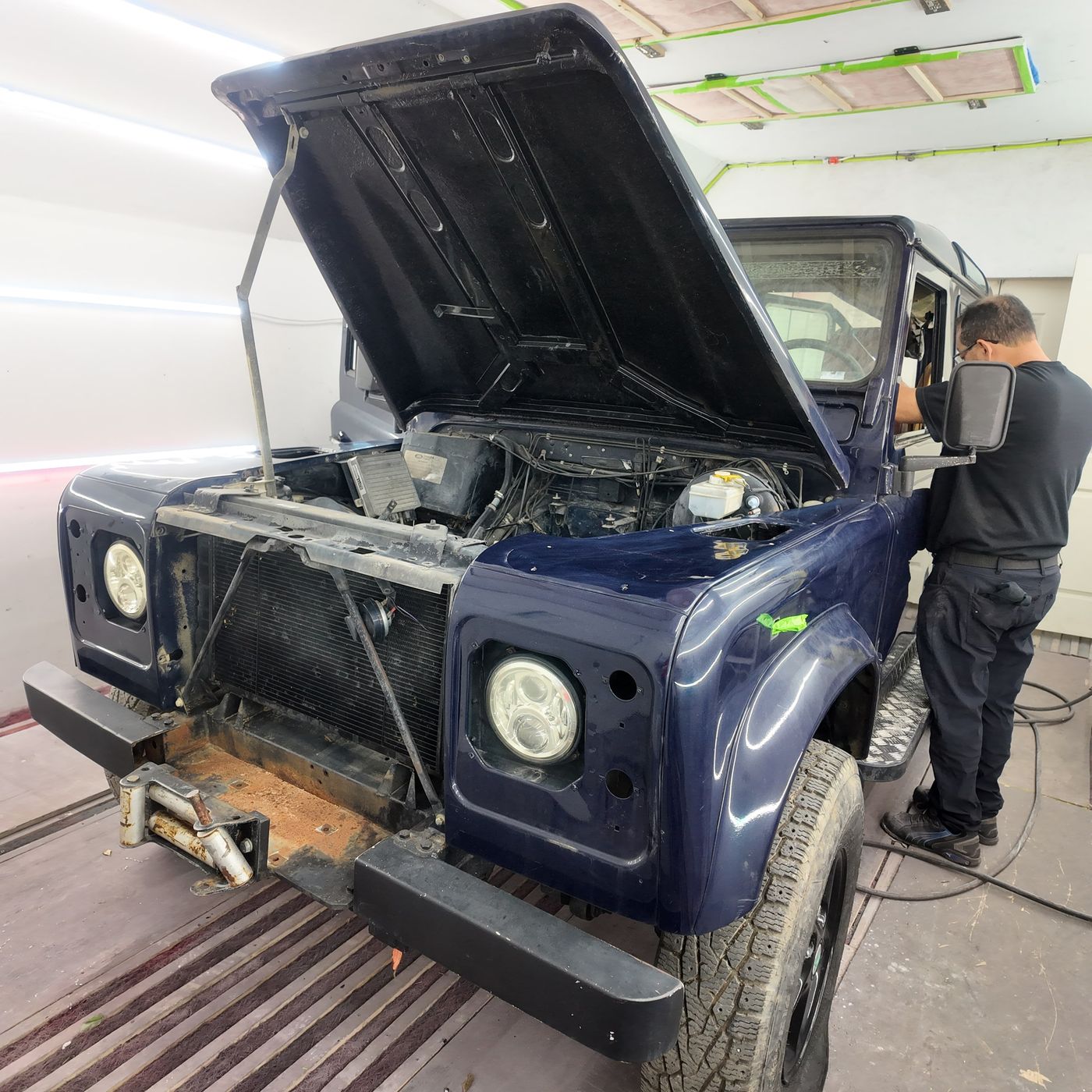 Dark blue Land Rover Defender in the shop with the hood up and front grille panel removed, technician working at the fender